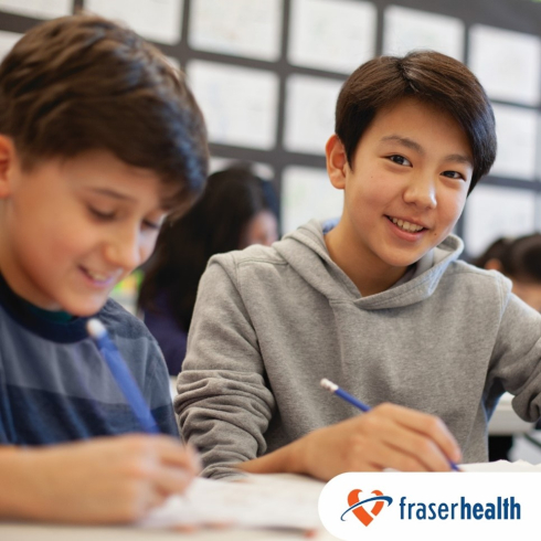 Two smiling boys work at a desk in a classroom.