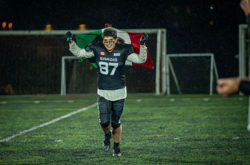 A student in football gear runs down a field holding a green, white and red flag above their head. 