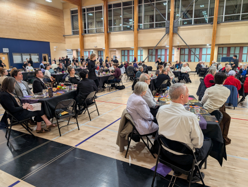 A large gym is filled with tables of people. Students in black serve meals. 