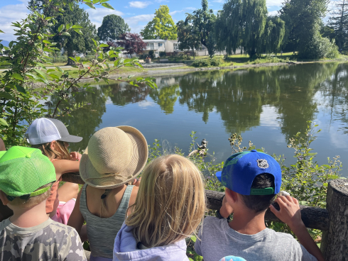 Children gather together and are looking out over a pond. It's sunny. Most are wearing hats. 