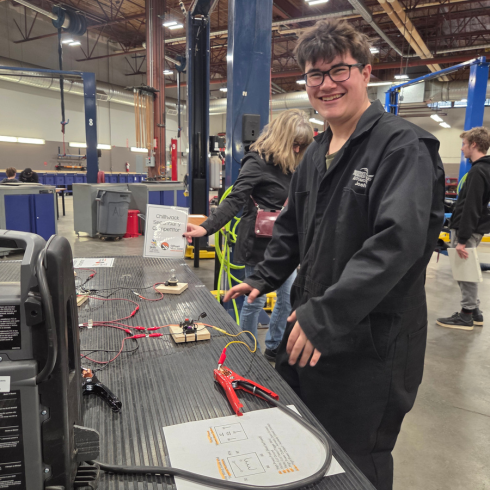 A student wearing a mechanic's suit smiles at the camera. On the surface in front of them are jumper cables and wires. 