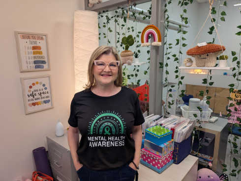 A woman with a blonde bob is smiling. She wears cat eye glasses and a black t-shirt that reads Mental Health Awareness. 