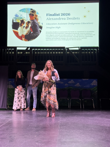 A woman with long hair and a long flowing dress smiles while holding a bouquet of flowers and a plaque. The screen above her reads: Alexandrea Desilets, Education Assistant. 