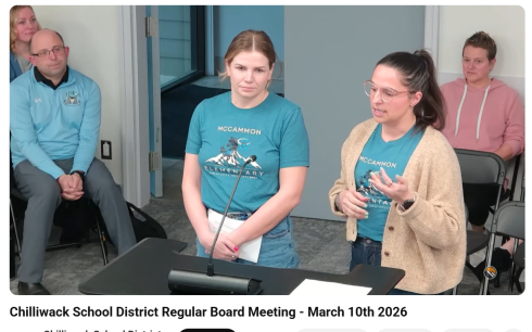 Two women in blue shirts stand at a podium. One wears a brown cardigan. They are speaking into a microphone. 
