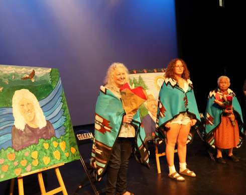 Two Elders and a student stand next to two portraits. They all have blue blankets draped over their shoulders. 