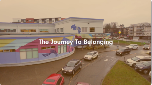 A screenshot of a video. The colour is muted. The words The Journey to Belonging sit overtop a still of school with a colourful mural. 