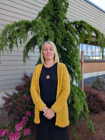 A woman wearing a black dress and yellow cardigan stands in front of an evergreen tree. She's smiling and has should length blonde hair. 