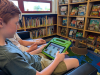 A boy sits and holds a tablet in a library. 