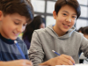 Two smiling boys work at a desk in a classroom.