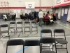 Chairs in forefront. Adults gathered at tables in a gym in the background.