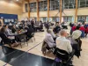 A large gym is filled with tables of people. Students in black serve meals. 