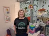 A woman with a blonde bob is smiling. She wears cat eye glasses and a black t-shirt that reads Mental Health Awareness. 