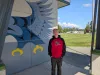 A man in a red and black hoodie stands in front of a building with a mural. Children play on a field in the background. 