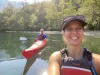 A woman in a life jacket sits in a kayak in the water. She is wearing and hat and has a big smile. There is a man in a kayak behind her who is also smiling.