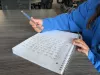 A student's hands and arms are visible as they write with a pencil in a white notebook.